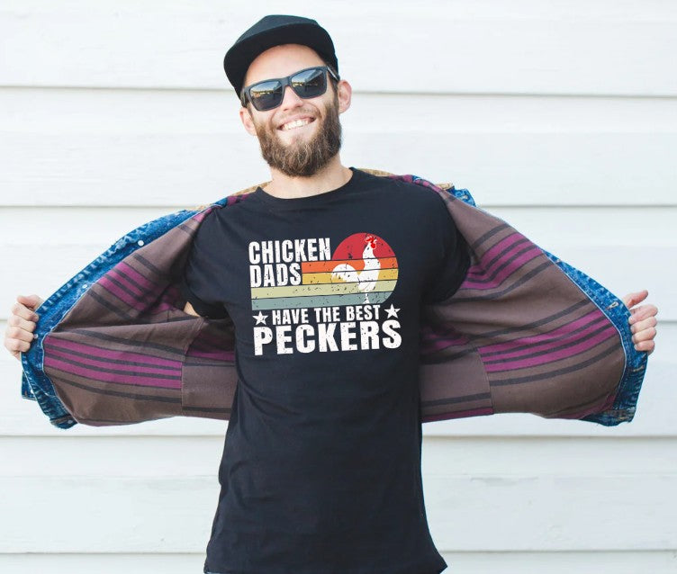 Man wearing a black t-shirt with 'Chicken dads have the best peckers' text and a graphic of a chicken, standing against a white wall.