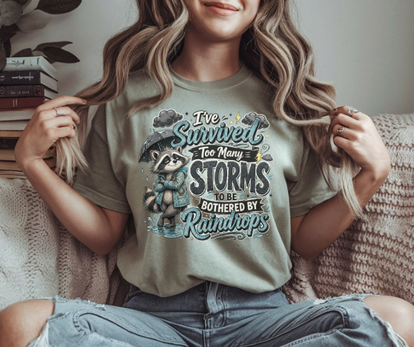 Funny shirt featuring I've Survived Too Many Storms to Be Bothered by Raindrops with a raccoon in a raincoat